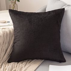 a black pillow sitting on top of a gray couch next to a white blanket and a vase with flowers