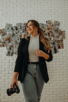 a woman standing in front of a white brick wall holding a camera and smiling at the camera