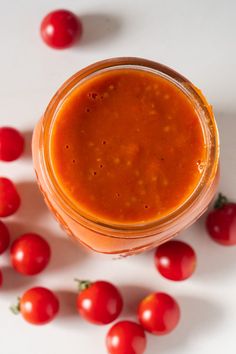 a glass jar filled with tomato sauce surrounded by small red tomatoes on a white surface