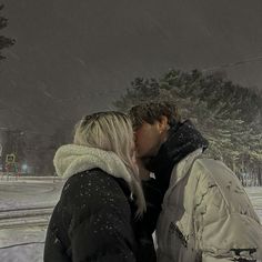 a man and woman kissing in the snow on a snowy night with trees behind them