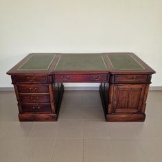 an antique desk with two drawers and green leather top, on tile flooring in front of a white wall