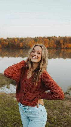 a woman standing in front of a body of water wearing jeans and a red sweater