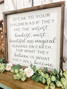 a framed sign with flowers and greenery on top of a wooden table in front of a white wall