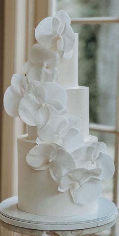 a three tiered white wedding cake with flowers on the top and bottom, sitting in front of a window