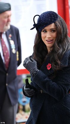 a woman in a black coat and hat with her hand on her hip, standing next to an older man