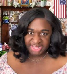 a woman with black hair and an american flag on the wall behind her is looking at the camera