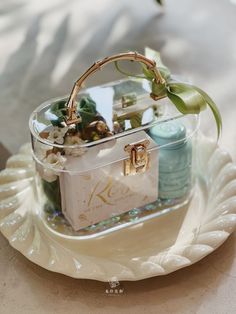 a clear basket with flowers and personal items in it on a white plate next to a candle