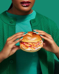 a woman is holding a large sandwich in her hands while wearing green sweater and blue nail polish