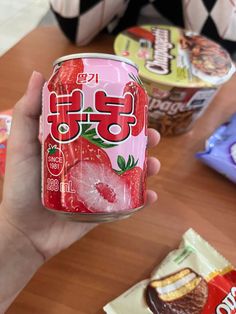 a person holding up a can of soda on top of a wooden table next to other snacks