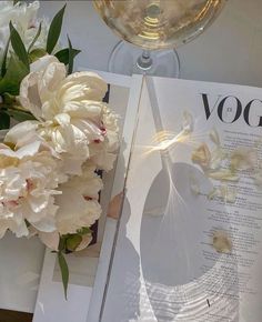 a glass of wine sitting next to a book with flowers on it and a vase filled with white peonies