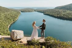 a bride and groom holding hands on top of a mountain overlooking a lake with mountains in the background