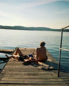 two people are sitting on a dock by the water and one is reading a book