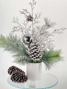 a glass table topped with a white vase filled with pine cones and evergreen branches next to a silver ornament