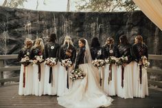 a group of women standing next to each other in front of a stone wall holding bouquets