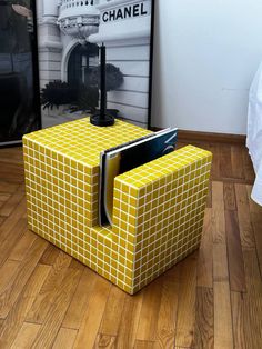 a yellow cube shaped end table with a magazine holder on it's side in front of a poster