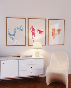 a white chair sitting next to a lamp on top of a wooden floor in front of two framed pictures