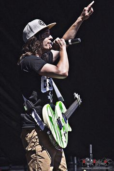 a man with long hair holding a guitar and pointing at something in the air while wearing a hat