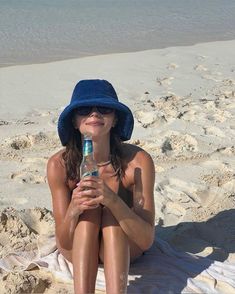 a woman in a blue hat is sitting on the beach and drinking a water bottle