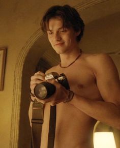 a shirtless young man holding a camera in his right hand and looking into the mirror