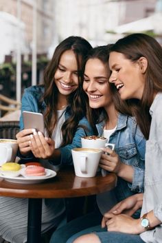 three young women sitting at a table looking at a cell phone and having a conversation