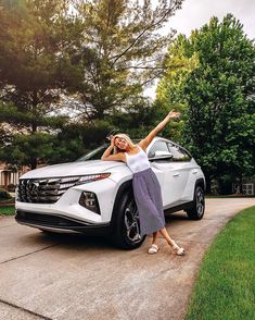 a woman leaning on the hood of a white car in front of trees and grass