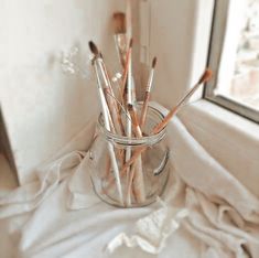 a glass jar filled with lots of paint brushes on top of a white cloth next to a window