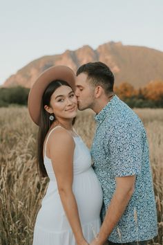 a pregnant couple kissing in the middle of a field with mountains in the back ground