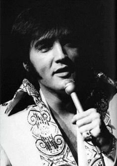 a black and white photo of a man holding a microphone in his right hand while wearing an elvis jacket