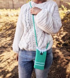 a woman wearing a white sweater and green crochet purse standing in front of a tree