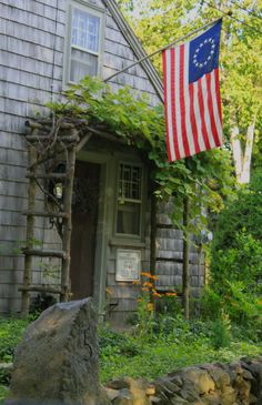 an american flag hanging from the side of a house next to a rock and tree