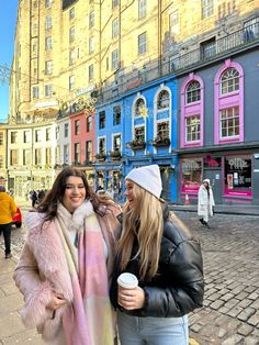 two women standing next to each other in front of colorful buildings and holding coffee cups