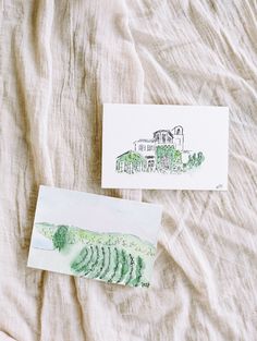 two cards are laying on a bed with white linens and one has a drawing of a house