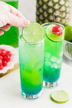 two glasses filled with green and blue liquid next to a pineapple on the side