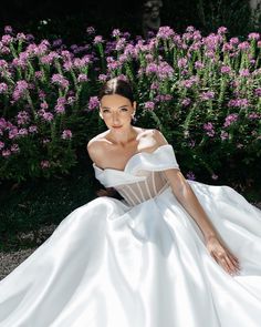 a woman in a white dress sitting on the ground next to purple flowers and bushes