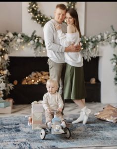 a man and woman are standing in front of a christmas tree with a baby on a tricycle