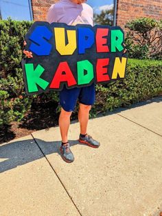 a man holding a sign that says super kaden in front of a building with bushes