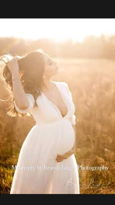 a pregnant woman in a white dress standing in a field with her hands on her head