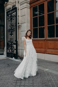 a woman in a white dress is standing on the sidewalk near an old building and looking at the camera