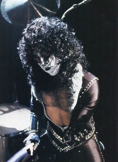 a man with long hair and makeup is dressed in black leathers, holding a drum