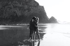 black and white photograph of two people hugging on the beach with mountains in the background