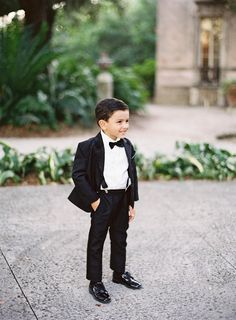 a young boy in a tuxedo and bow tie standing on the sidewalk with his hands in his pockets