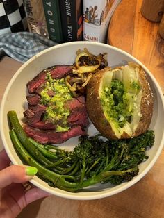 a person holding a bowl with steak, broccoli and potatoes in it on a table