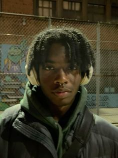 a young man with dreadlocks standing in front of a chain link fence and building