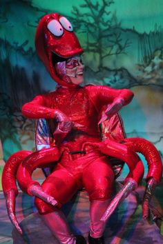 a man dressed in red sitting on top of a chair with a giant spider costume over his head