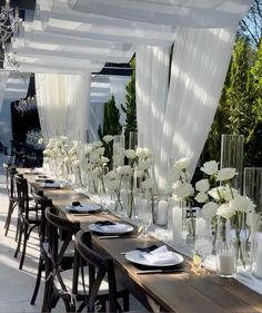 a long table with white flowers in vases and plates on the tables under an awning