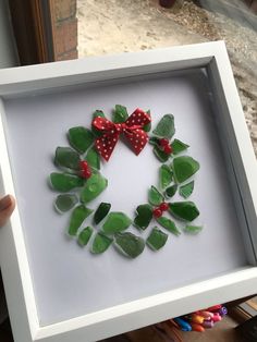 a white frame holding a green and red wreath made out of sea glass with bows