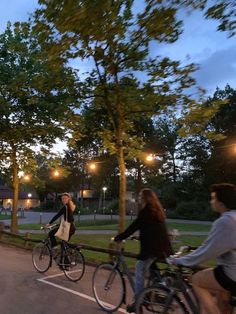 three people riding bikes in a parking lot at night with street lights on the trees