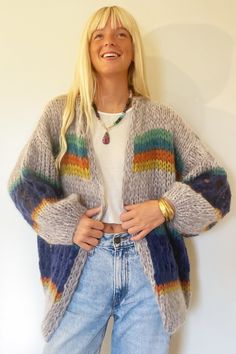 a woman standing in front of a white wall wearing a colorful cardigan sweater and jeans