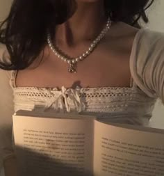 a woman holding an open book in front of her face and wearing a pearl necklace