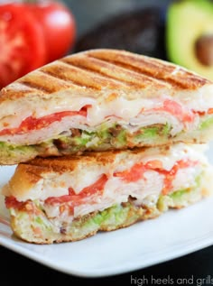 a grilled sandwich on a white plate with an avocado and tomato in the background